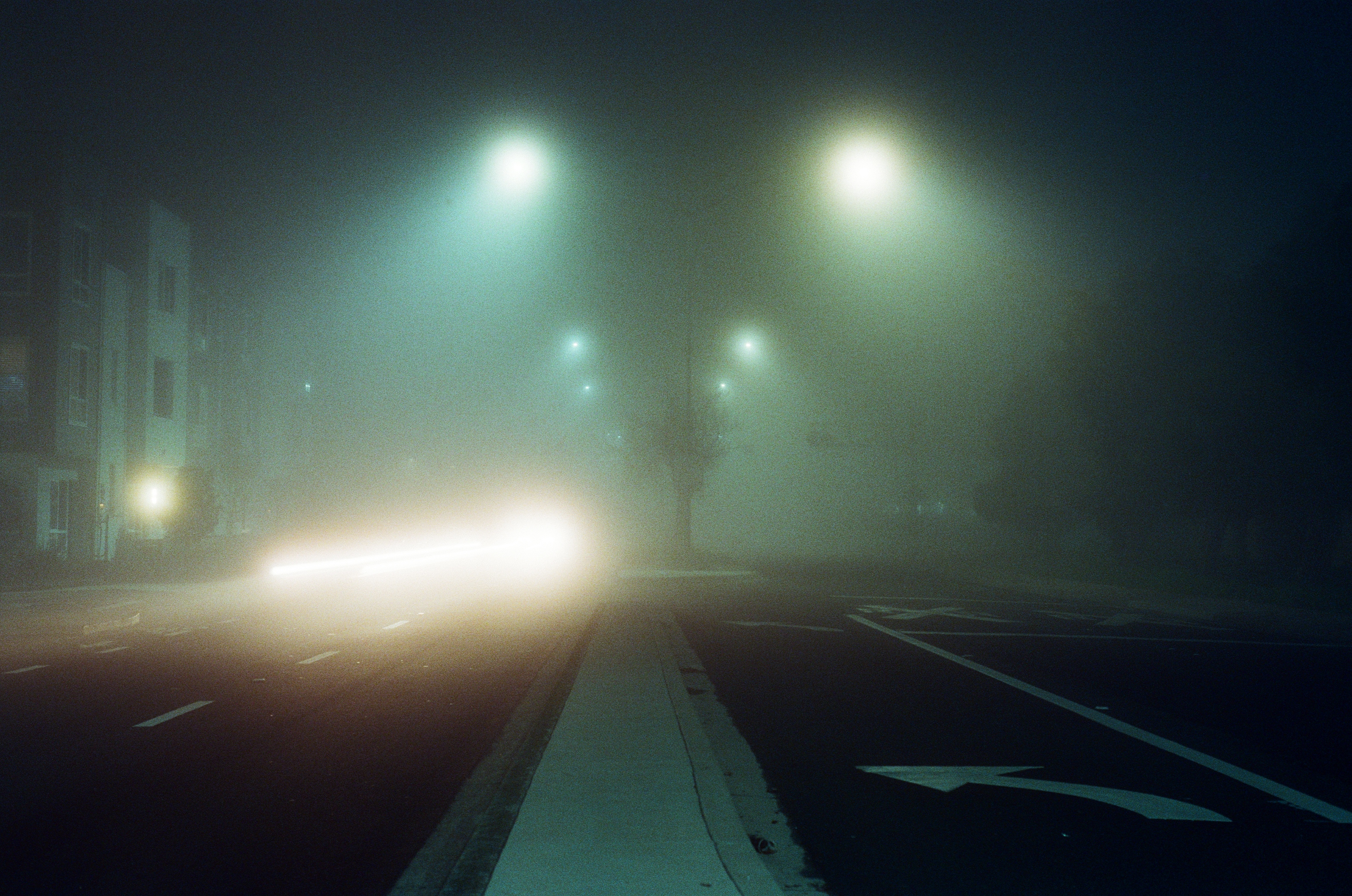 Foggy street at night, streetlights glowing through thick mist, car headlights in the distance